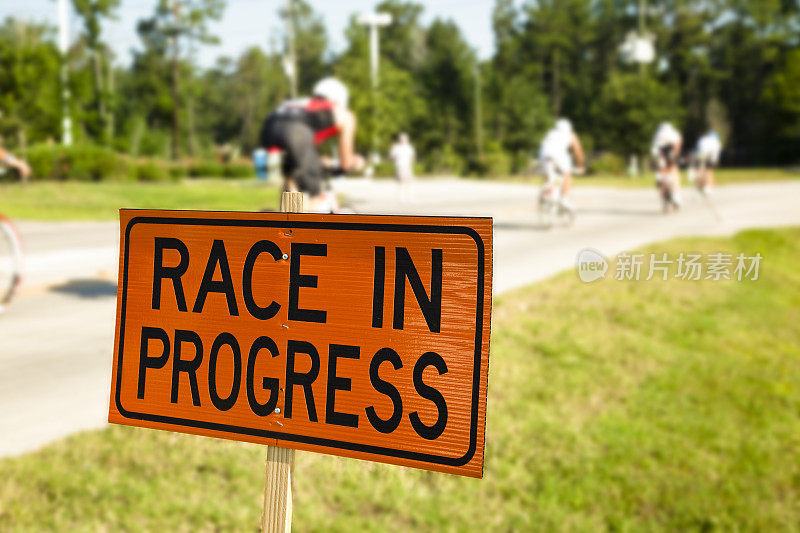 一大群骑自行车的人在比赛。“Race in Progress”标志着前景。